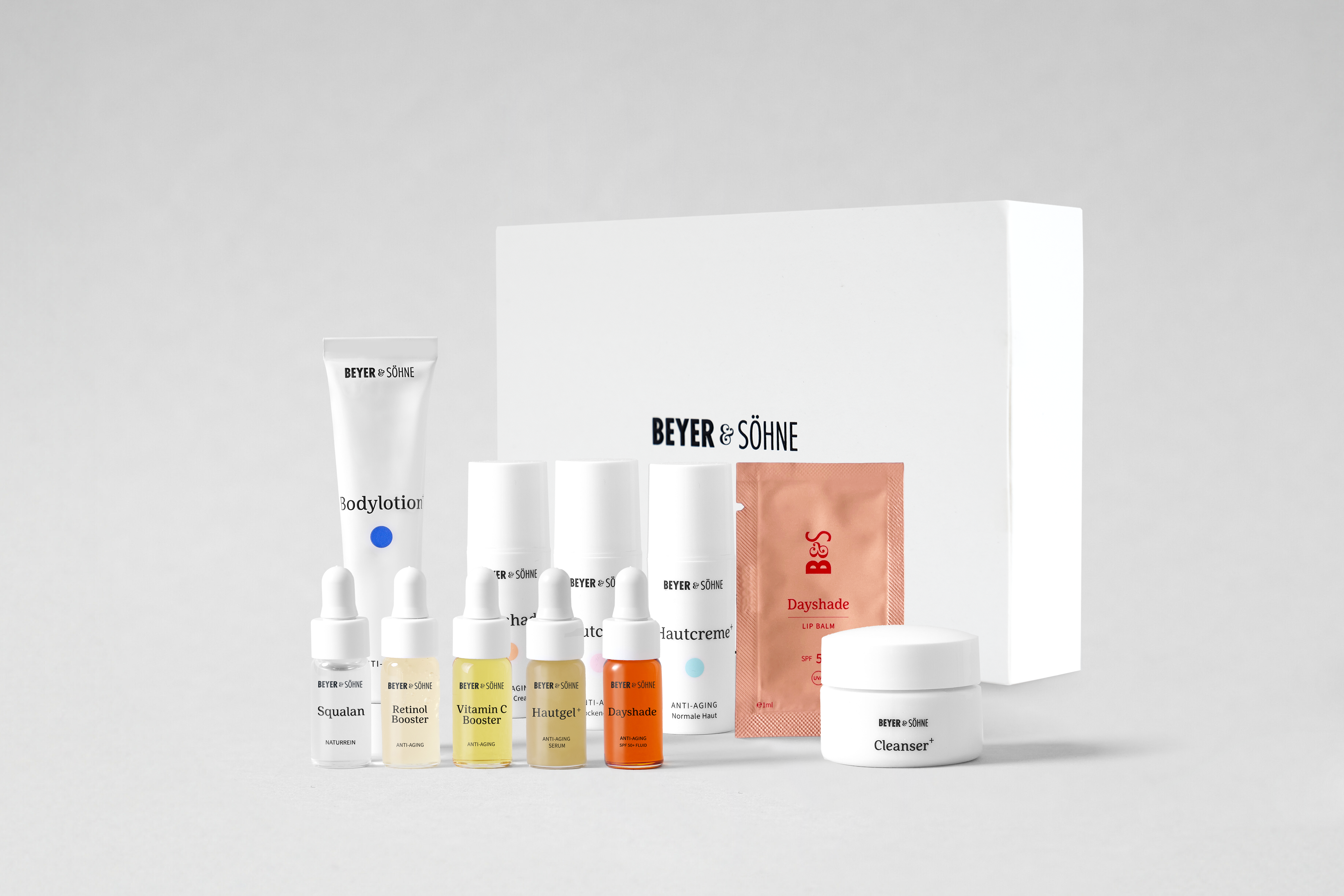 Testerset von BEYER & SÖHNE mit verschiedenen Hautpflegeprodukten als niedliche Minigrößen, darunter Bodylotion, Cleanser, drei Booster (Retinol, Vitamin C, Squalan), zwei Hautcremes, ein Dayshade Lip Balm, ein Sachet mit Dayshade Lippenpflege sowie eine weiße Produktbox im Hintergrund mit BEYER & SÖHNE Logo